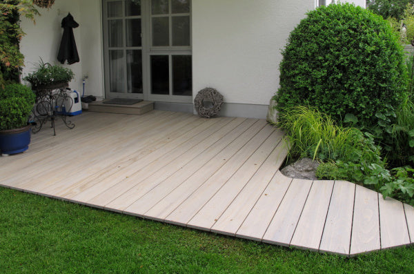 Sicht auf eine stilvolle Holzterrasse aus Acoya Holz, die in einen gepflegeten Garten integriert ist, mit Weiß- und Grüntönen und einer gemütlichen Sitzgelegenheit.