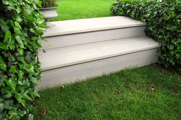 Drei Stufen aus hellem Acoya Holz führen in einen grünen Garten, umgeben von Efeu und einem blühenden Gehölz, perfekt für einen eleganten Außenbereich.